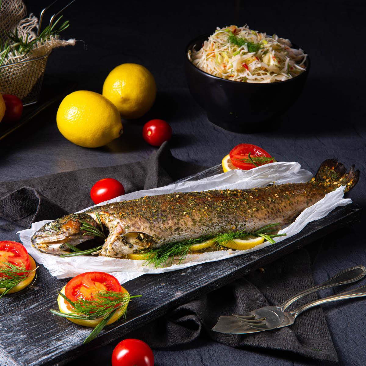 Gebackene Forelle mit frischen Kräutern und Gemüsegarnitur Gebackene Forelle auf einem Tablett, dekoriert mit Zitronenscheiben, Tomaten und frischen Kräutern, im Hintergrund eine Schüssel mit Krautsalat und Zitronen