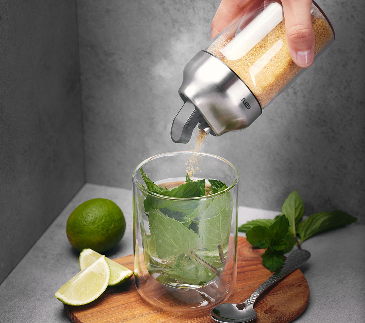 Zuckerstreuer beim Zubereiten eines frischen Getränks mit Limette und Minze Hand hält Zuckerstreuer über einem Glas mit Minzblättern, Limettenstücken und einem Holzbrett im Hintergrund