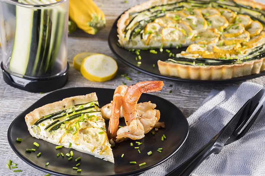 Zucchini-Zitronentarte mit gebratenen Riesengarnelen Ein Stück Gemüsequiche mit Zucchini und Garnelen auf einem schwarzen Teller, daneben ein Spiralschneider und eine gelbe Zucchini.
