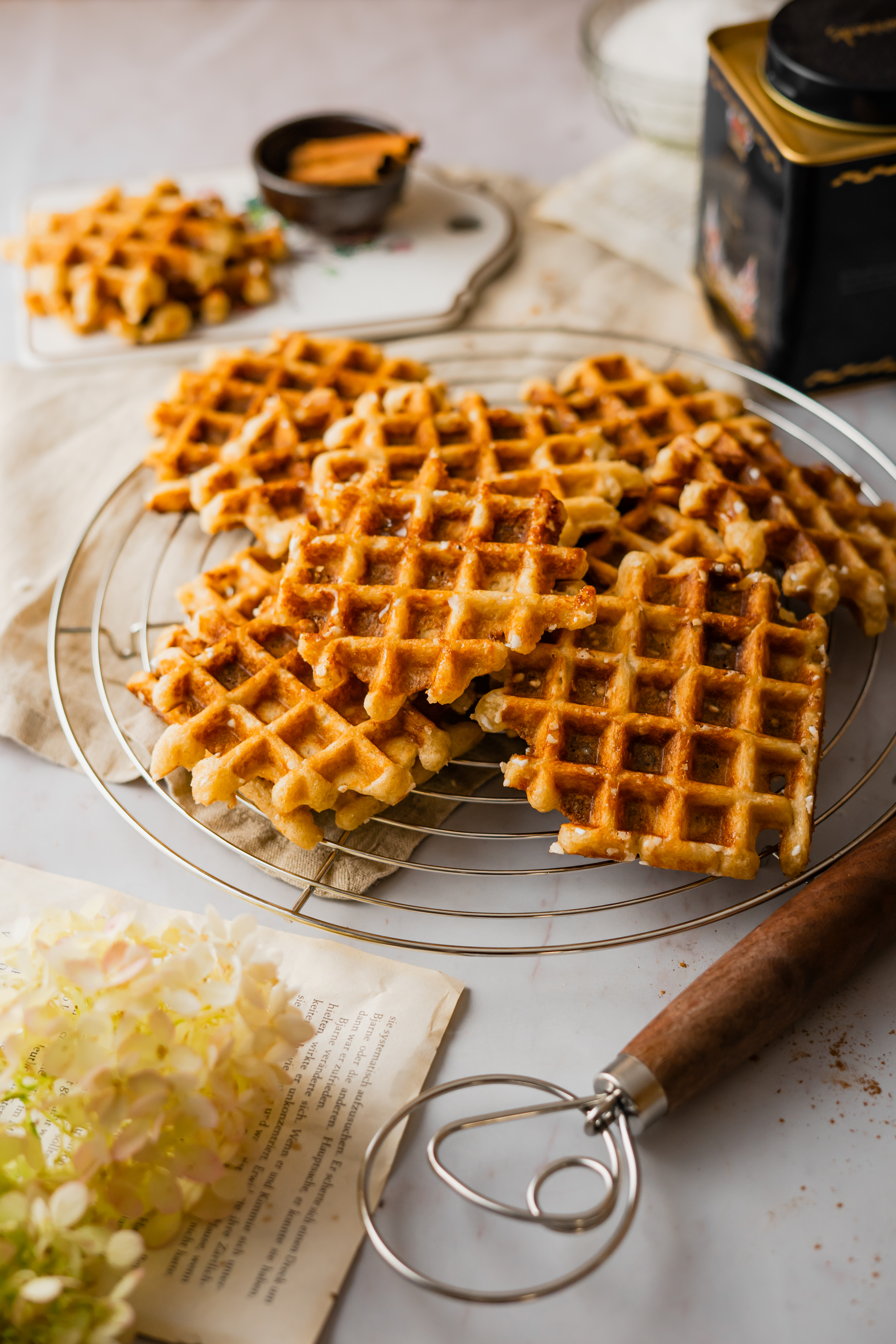 Karamellisierte Belgische Waffeln  Goldbraune belgische Waffeln auf einem runden Abkühlgitter, umgeben von Zimtstangen und Blumen