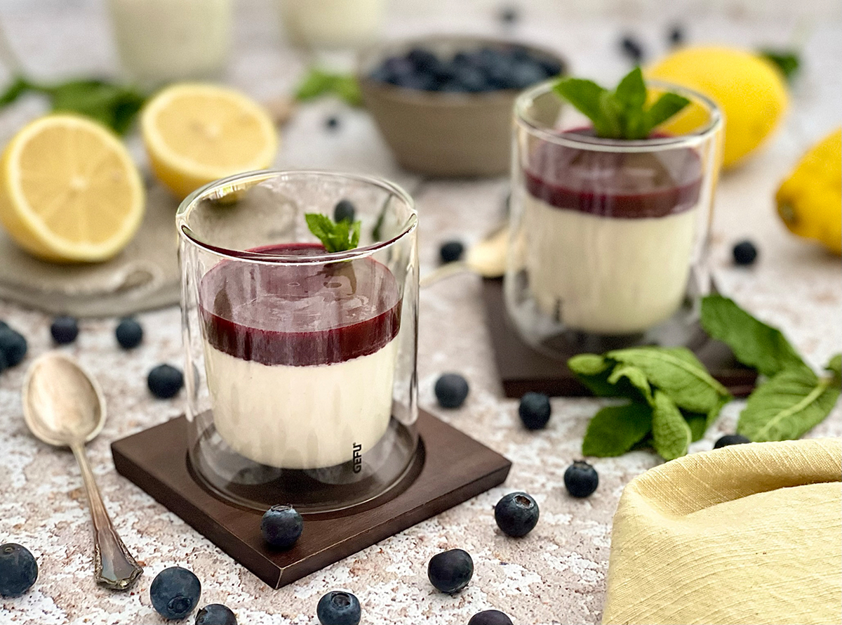 Glas mit Schichtdessert aus Joghurt und Fruchtsauce auf Holzteller Ein Dessert im Glas mit weißer Joghurtcreme und roter Fruchtsauce, dekoriert mit Minzblättern, umgeben von Zitronen und Heidelbeeren
