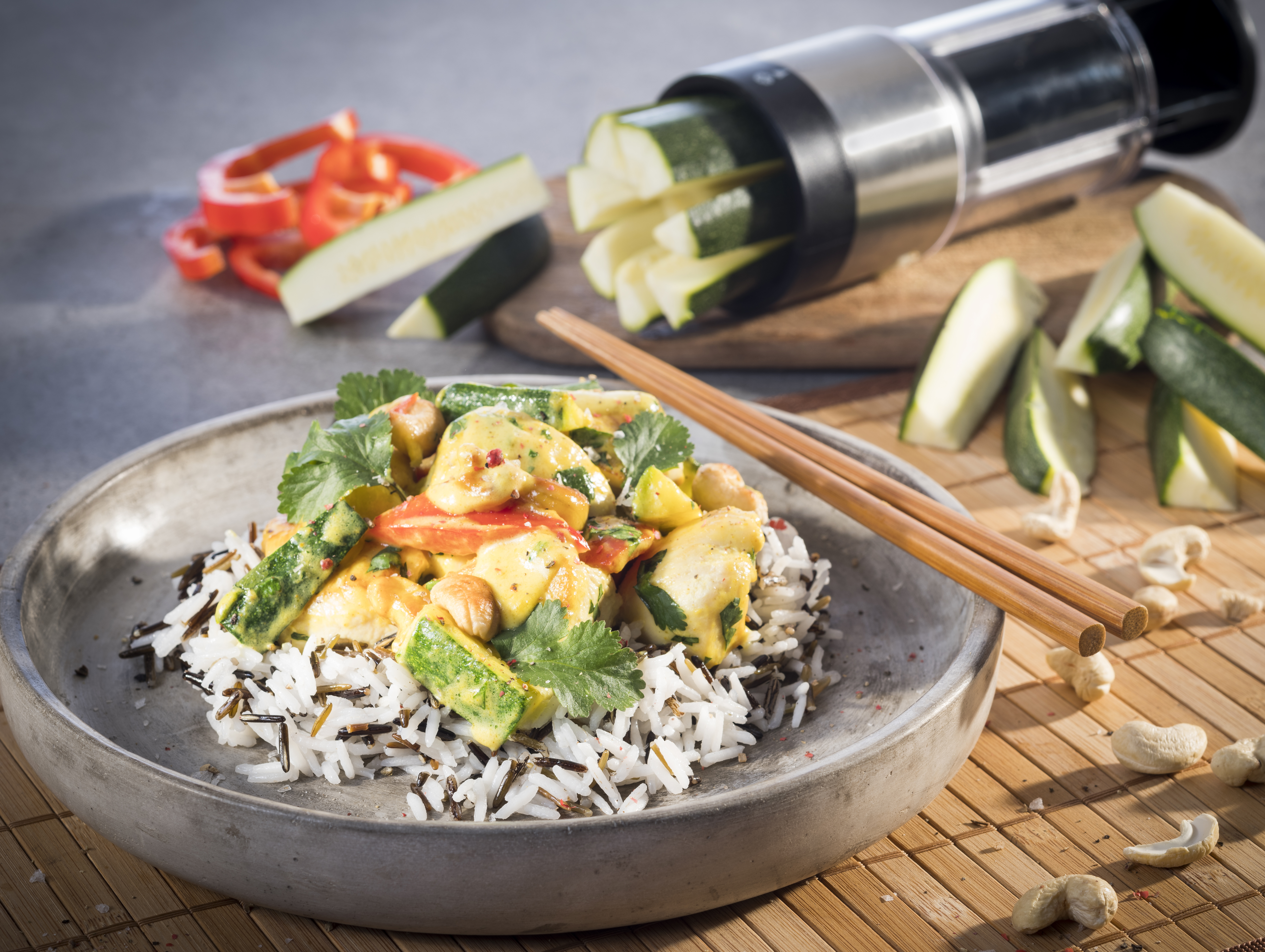 Hähnchencurry mit Zucchini, Paprika, Cashewkernen und Basmatireis Nahaufnahme von asiatischem Gemüse-Curry mit Reis und Cashewnüssen, dekoriert mit Koriander, serviert auf einem modernen Teller. Im Hintergrund geschnittenes Gemüse und ein Spiralizer.
