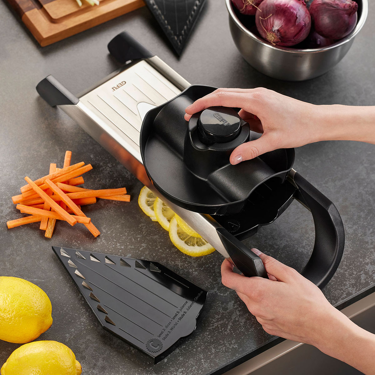Hochwertige Mandoline mit Sicherheitshalter, geeignet für präzises Schneiden von Gemüse Person bedient eine Mandoline mit Sicherheitshalter auf einer Küchenzeile, umgeben von geschnittenem Gemüse und Zitrone