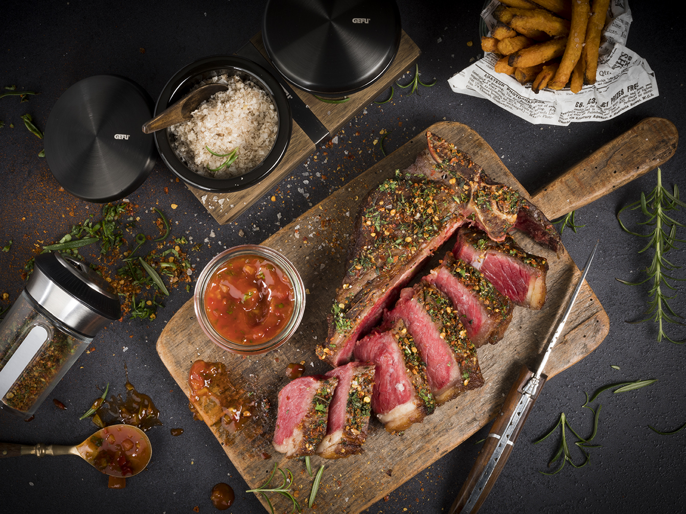 Porterhouse Steak mit einer Kräuter-Gewürz-Kruste, Salsa und Süßkartoffel-Pommes Ein perfekt gebratenes, in Scheiben geschnittenes Steak liegt auf einem Holzbrett, umgeben von Gewürzen, einer Schale mit roter Soße und einer Portion Pommes Frites.