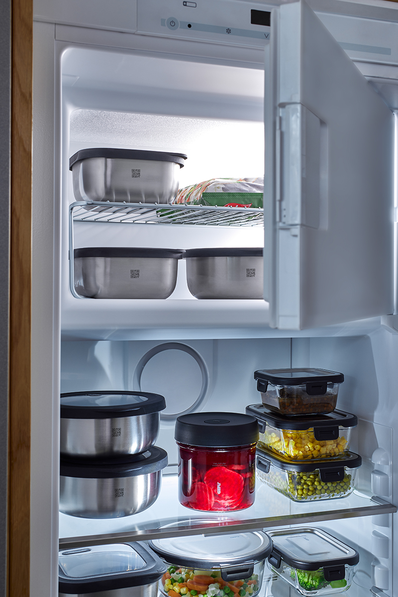 Organisierter Kühlschrank mit Vorratsbehältern Ein geöffneter Kühlschrank mit mehreren Vorratsbehältern aus Edelstahl und Glas, gefüllt mit verschiedenen Lebensmitteln.