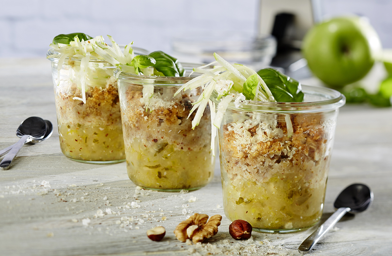 Knuspriges Bircher Apfel-Basilikum Crumble Drei Gläser mit Apfel-Crumble, garniert mit Streuseln und Basilikum, neben Nüssen und Löffeln auf einem Tisch