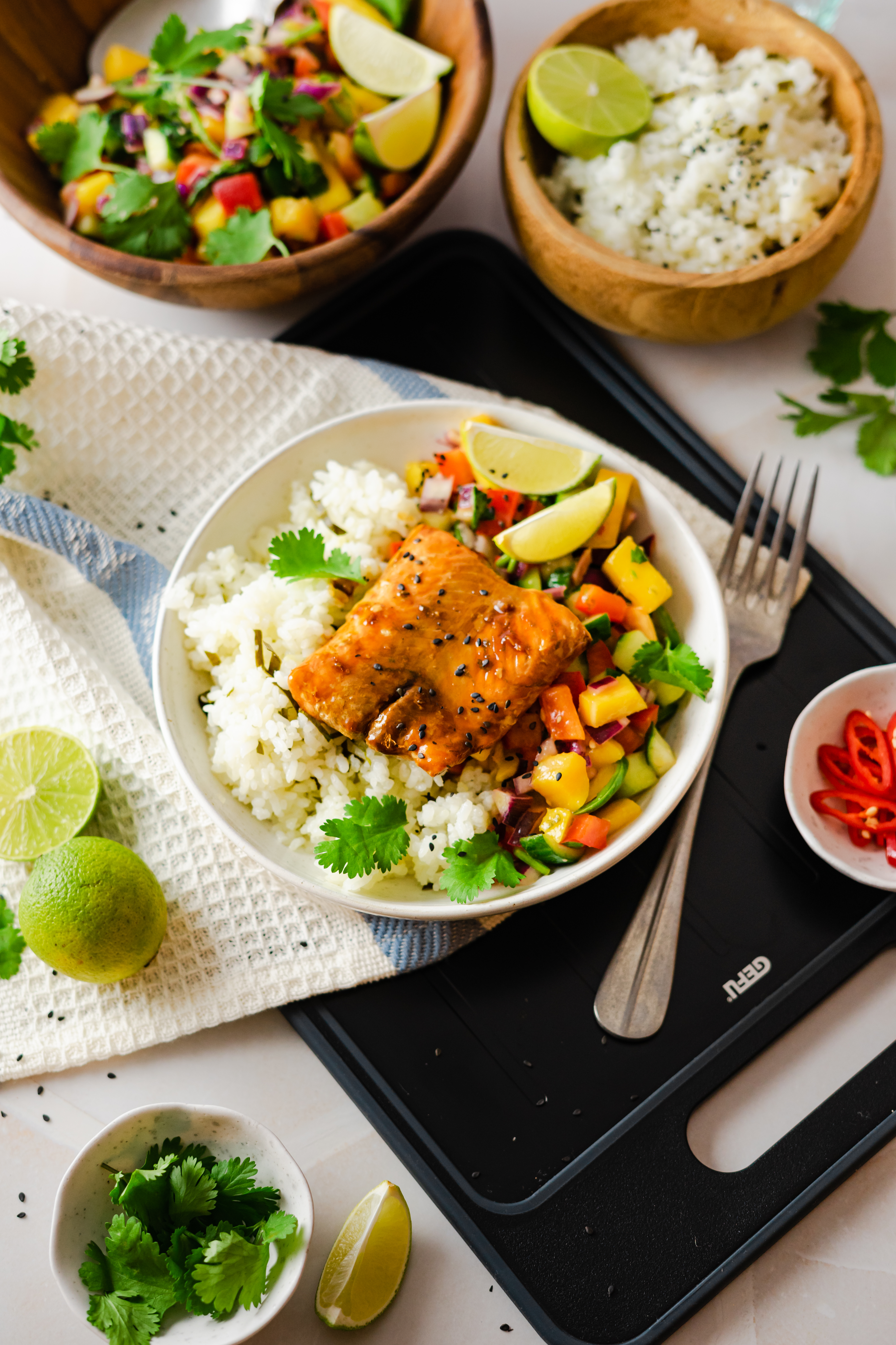 Lachs mit Mango Salsa und Limettenreis Eine Schüssel mit gegrilltem Lachs, serviert auf Reis mit bunter Gemüsesalsa und Limettenscheiben, umgeben von Kräutern und weiteren Beilagen.