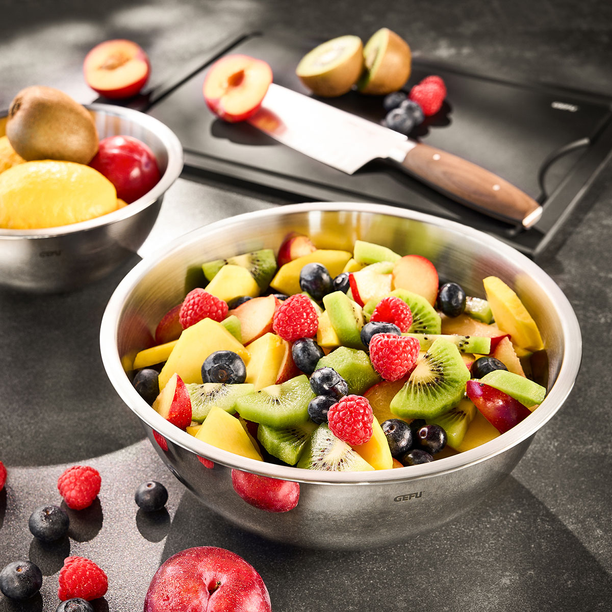 Frischer Obstsalat in einer Edelstahlschüssel mit Himbeeren, Blaubeeren, Kiwi und Mango Eine Edelstahlschüssel mit gemischtem Obstsalat aus Kiwi, Himbeeren, Blaubeeren und Mango auf einem Tisch mit weiteren Obstsorten und einem Schneidebrett im Hintergrund