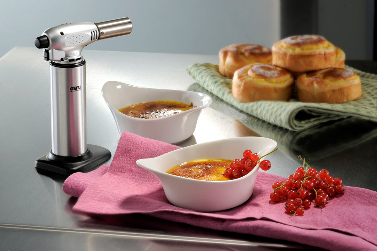 Küchenbrenner für perfekte Crème Brûlée Ein Küchenbrenner neben einer Crème Brûlée mit roten Johannisbeeren, im Hintergrund süße Gebäckstücke.