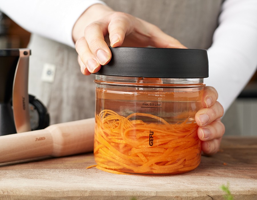 Bunte Fermentierte Möhrenspiralen  Person hält einen Glasbehälter mit Karottennudeln, verschlossen mit einem schwarzen Deckel, auf einem Holzbrett.