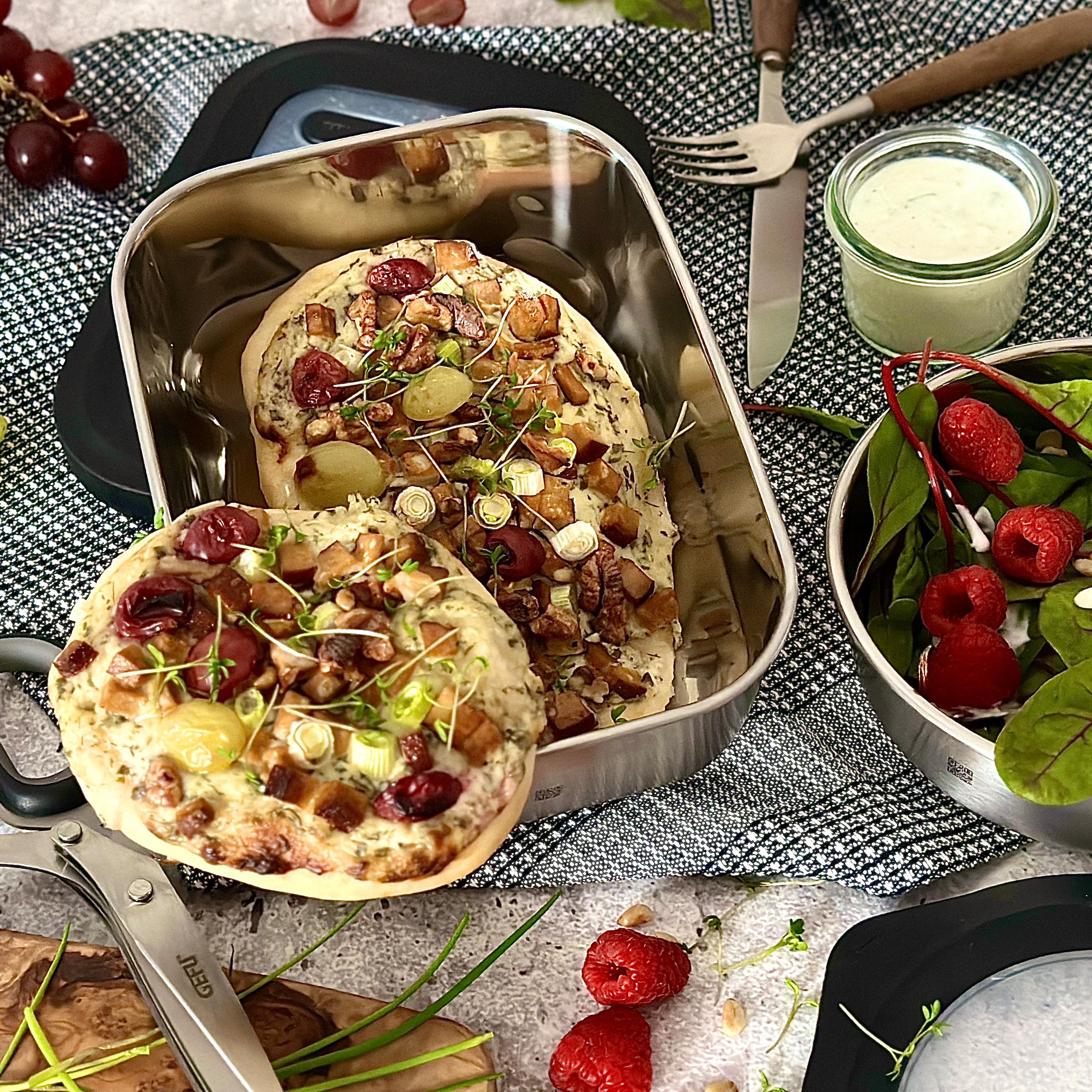 Meal Prep – vegane Flammkuchen und Salat Zwei Fladenbrote in einer Edelstahl-Lunchbox, garniert mit Trauben, Kirschen und Frühlingszwiebeln. Daneben ein Salat mit Himbeeren und ein Glas mit Dip.