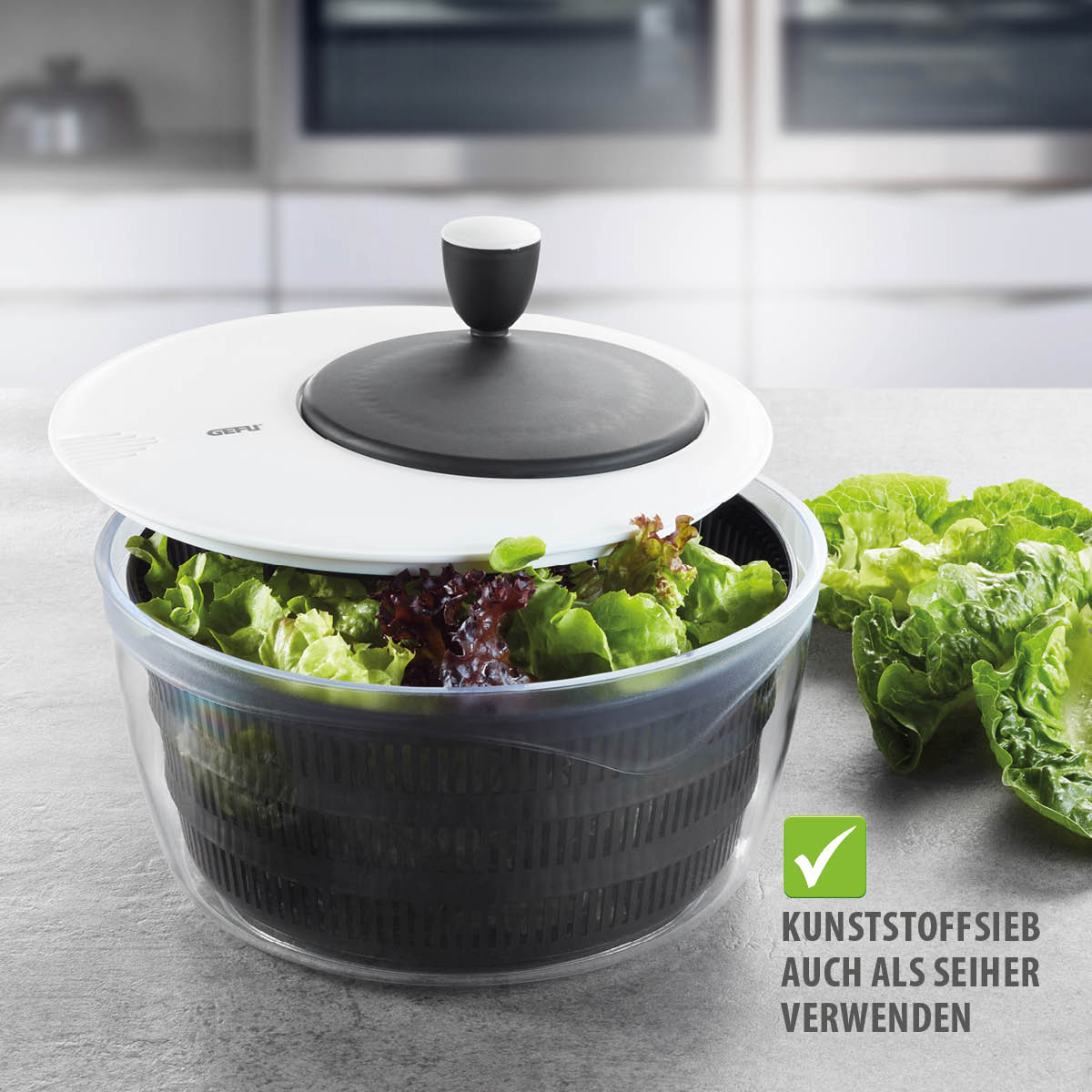 Hochwertige Salatschleuder mit Siebfunktion Kunststoff-Salatschleuder mit schwarzem Sieb und grünem Salat vor einer modernen Küche, kann auch als Sieb verwendet werden.