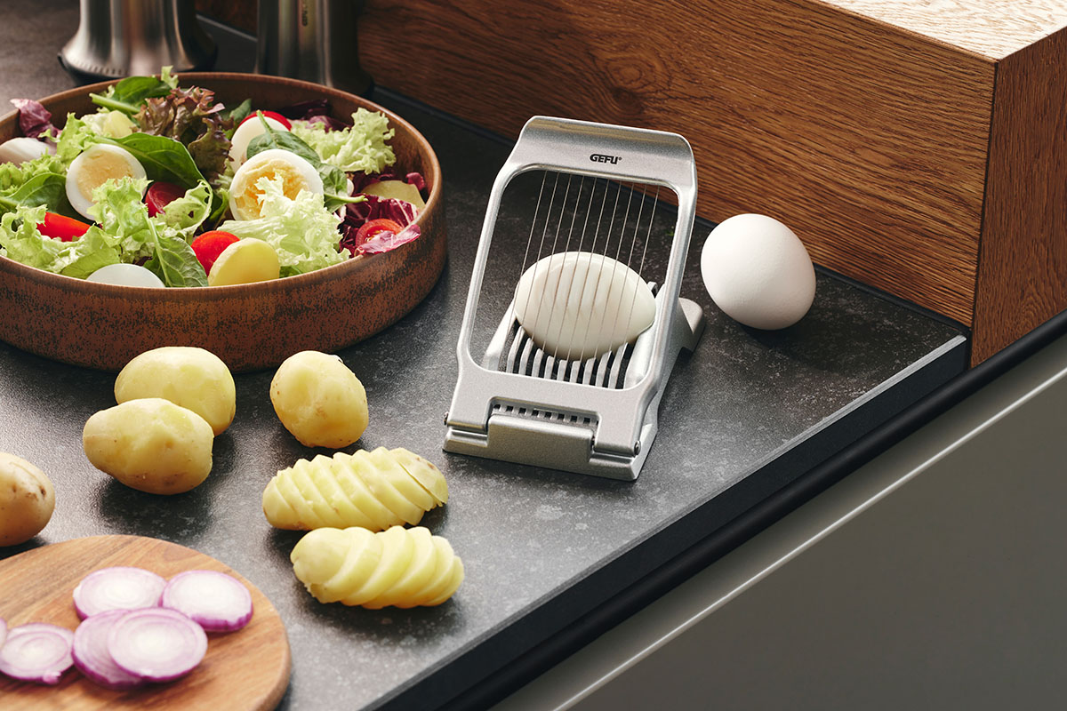 Ein Eierschneider auf einer Arbeitsplatte, daneben Salat, Eier und geschnittene Kartoffeln