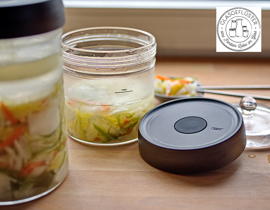 Weißes Kimchi Offene Fermentationsgläser mit geschnittenem Gemüse in Flüssigkeit auf einem Holztisch. Ein Glasdeckel liegt daneben. Logo von 'Glasgeflüster' im Hintergrund.