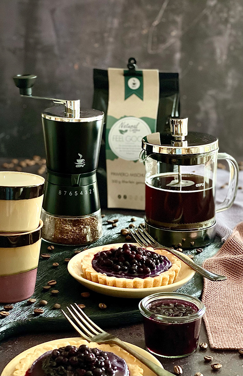 Ein gedeckter Tisch mit einer French Press, Kaffeebohnen, Heidelbeertörtchen und einer Kaffeemühle.