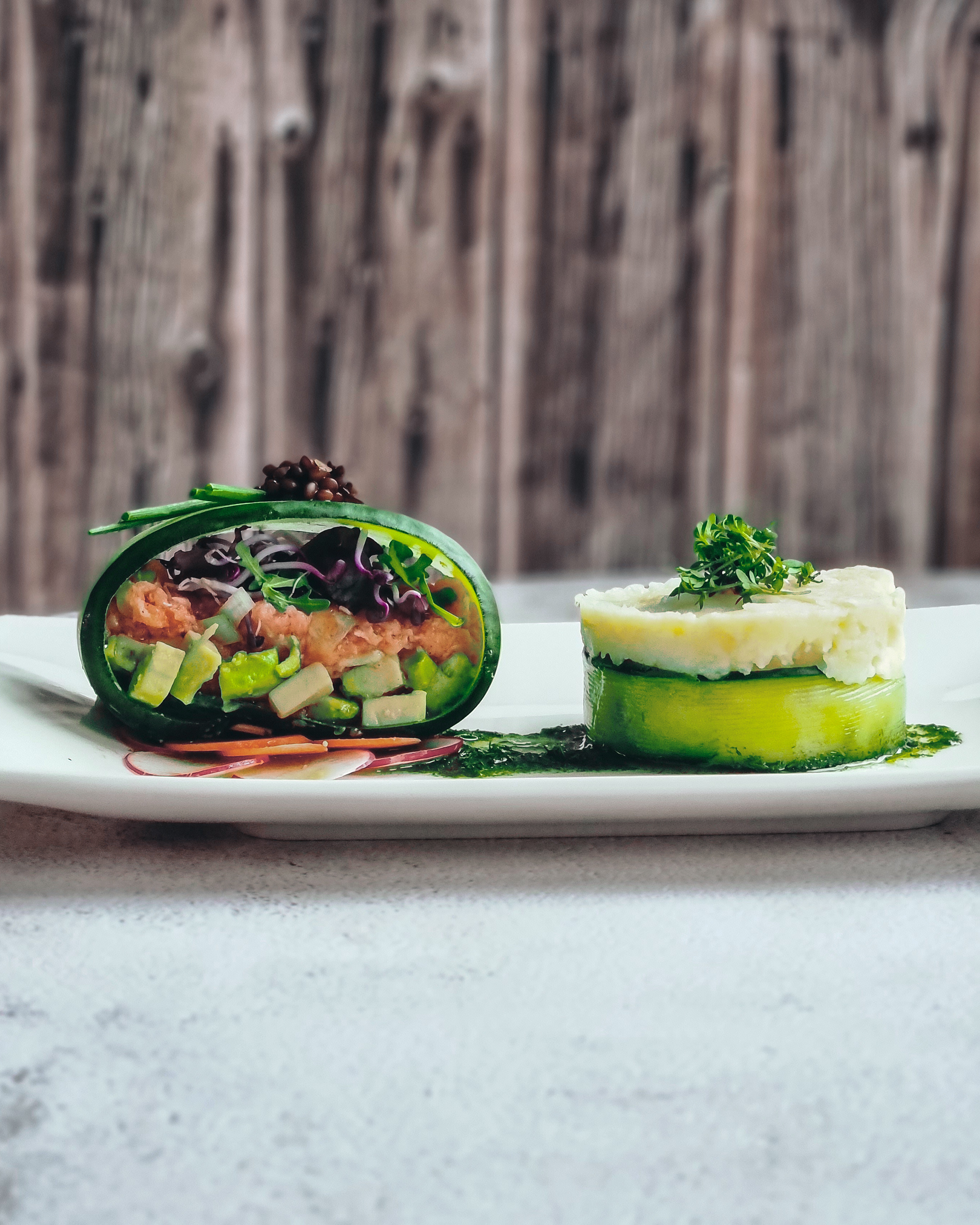 Mariniertes Lachstatar mit Beilage Eine kunstvoll angerichtete Gourmet-Vorspeise mit Avocado, buntem Gemüse und Kartoffelpüree auf einem weißen Teller. Im Hintergrund Holzwand.