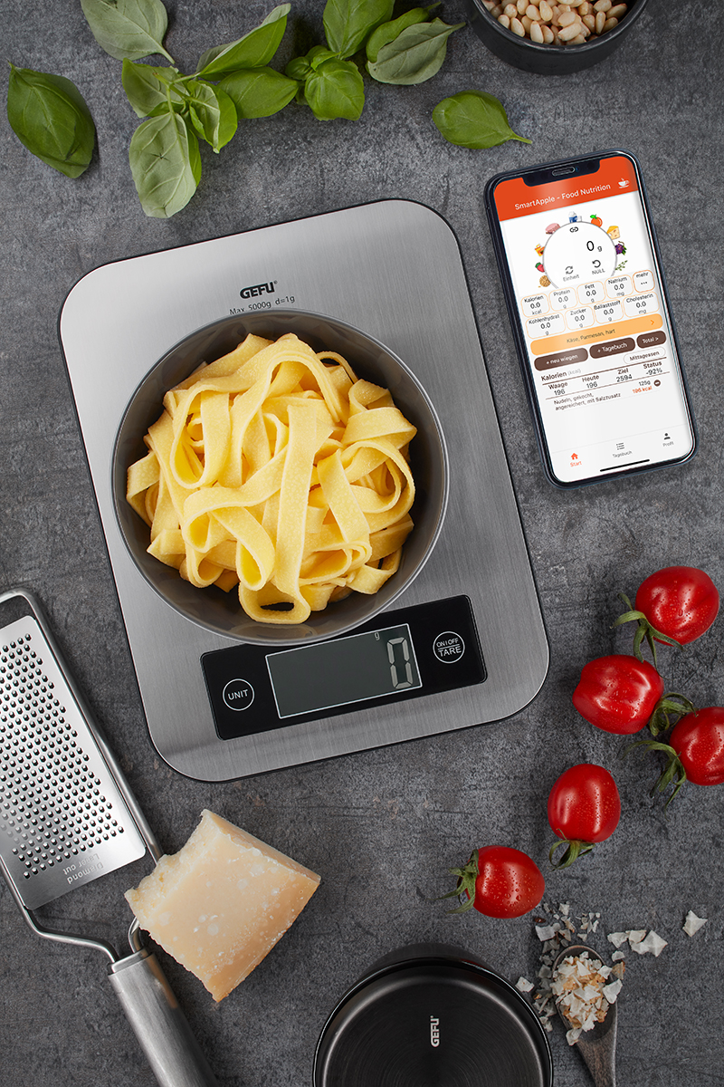 Küchenwaage mit frischen Zutaten und Smartphone-App Küchenwaage mit Nudeln und Smartphone-App, umgeben von frischen Zutaten wie Tomaten, Basilikum und Parmesan.