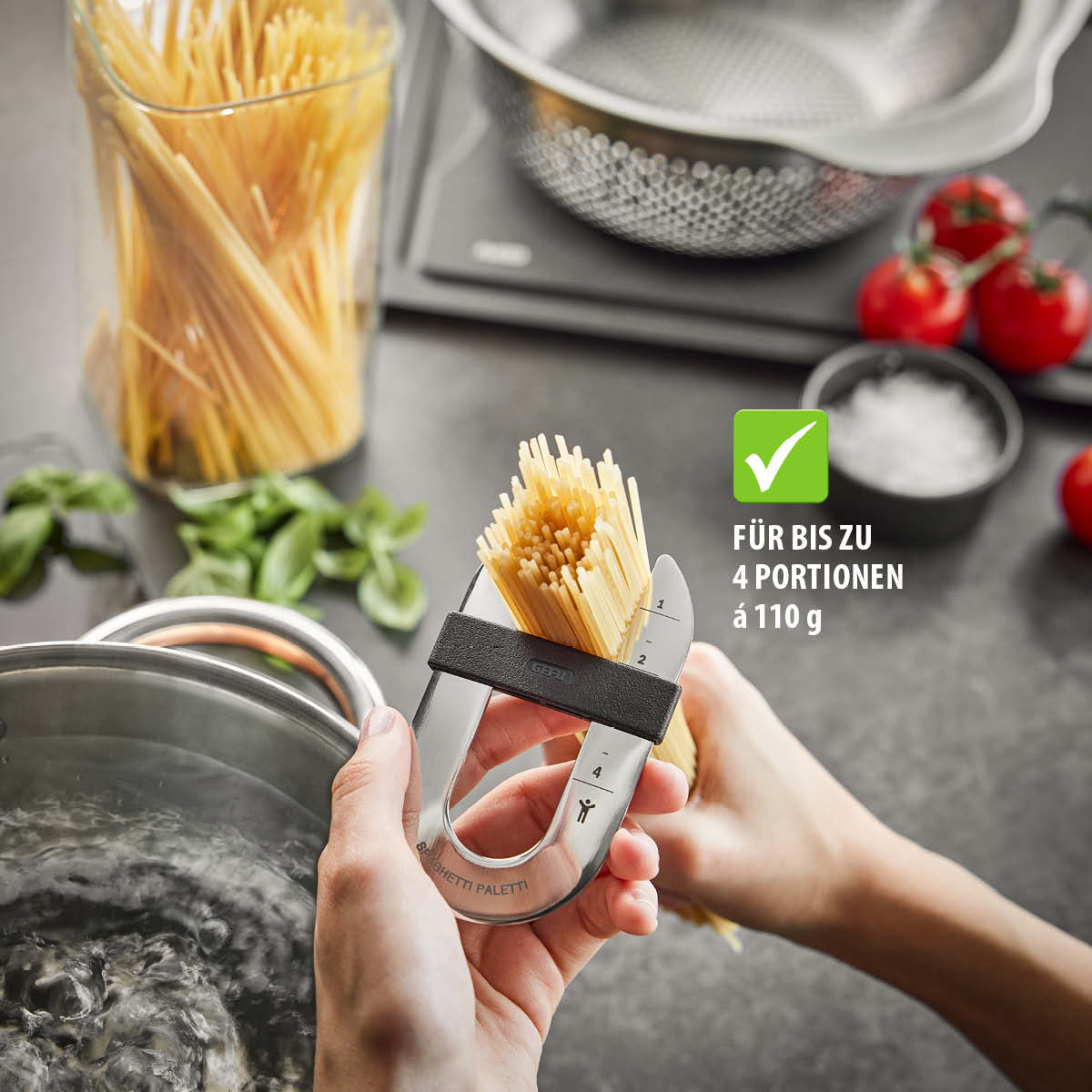 Spaghetti-Portionierer zeigt Portionen für bis zu 4 Portionen Spaghetti, je 110 g, über einem Kochtopf mit Basilikum und Tomaten im Hintergrund.