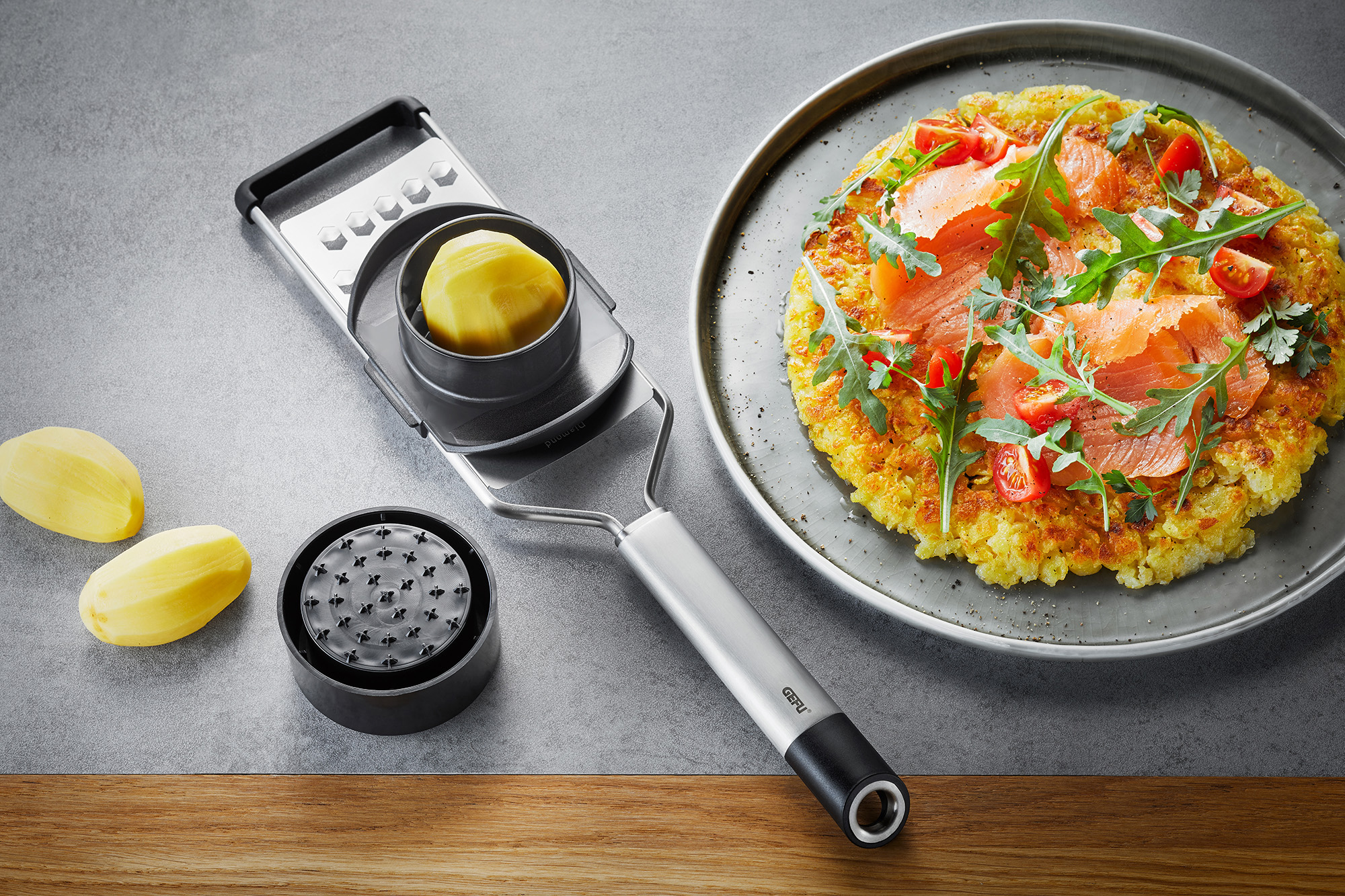 Kartoffelreibe mit leckerem Kartoffelpuffer und Räucherlachs Kartoffelreibe neben einem Teller mit Kartoffelpuffer, Räucherlachs, Rucola und Tomaten auf grauem Hintergrund