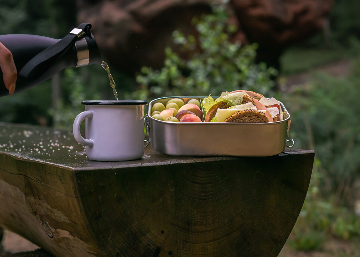 Picknick im Freien mit Thermoskanne und Lunchbox Eine Hand gießt Flüssigkeit aus einer Thermoskanne in eine weiße Tasse neben einer Lunchbox mit Brot, Käse, und Obst auf einem Holztisch im Freien.