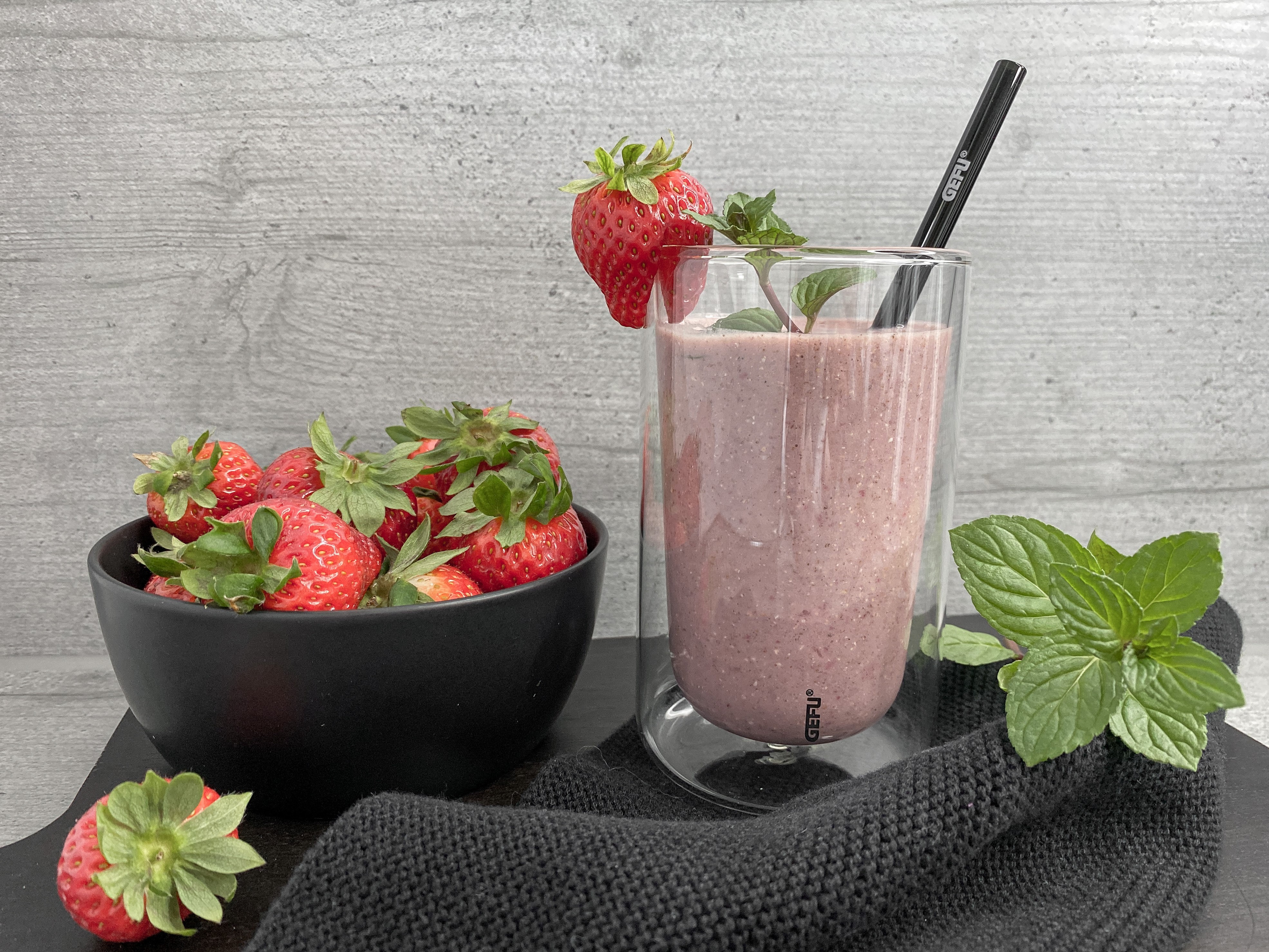 Erdbeer-Smoothie Ein Glas mit Erdbeer-Smoothie und Minzdekoration neben einer Schale voller frischer Erdbeeren, mit einem schwarzen Strohhalm und einem dekorativen Tuch.