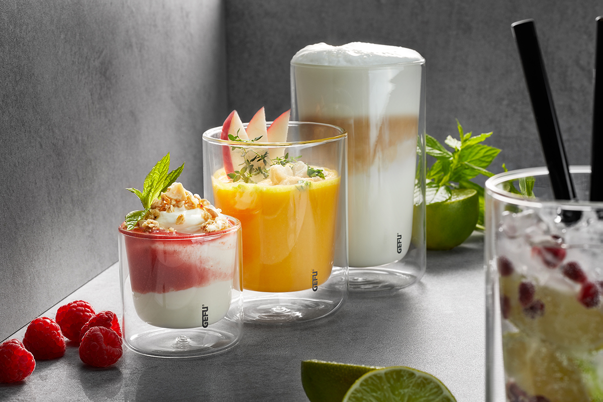 Doppelwandige Gläser mit verschiedenen Getränken und Desserts Drei doppelwandige Gläser mit einem Himbeer-Dessert, einem Mango-Drink und einem Latte Macchiato auf grauem Hintergrund, dekoriert mit frischen Himbeeren und Limetten.