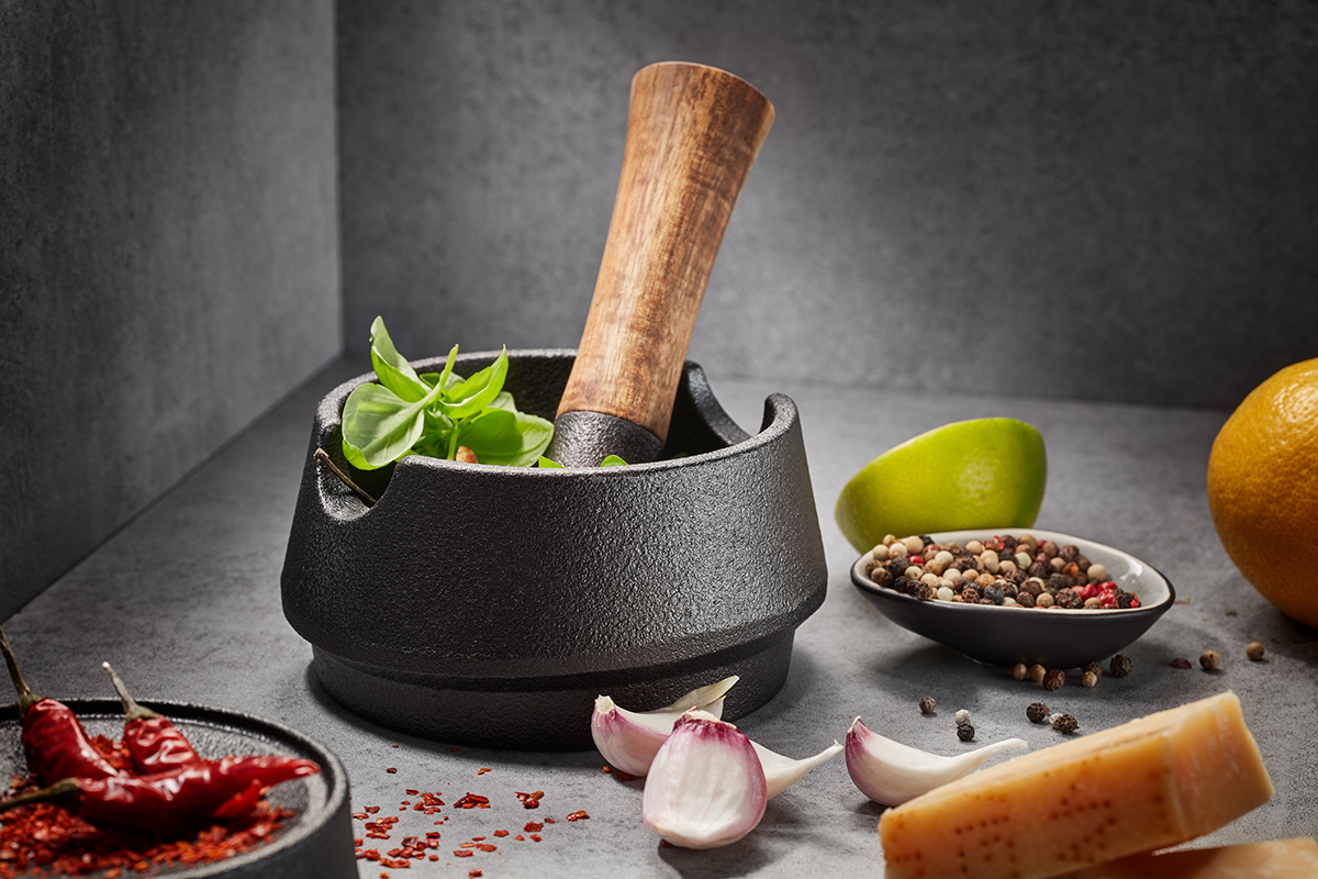 Hochwertiger Mörser aus Gusseisen mit Holzstößel für frische Kräuter Hochwertiger Mörser aus Gusseisen mit Holzstößel, umgeben von Kräutern, Gewürzen und Knoblauchzehen auf einem grauen Hintergrund