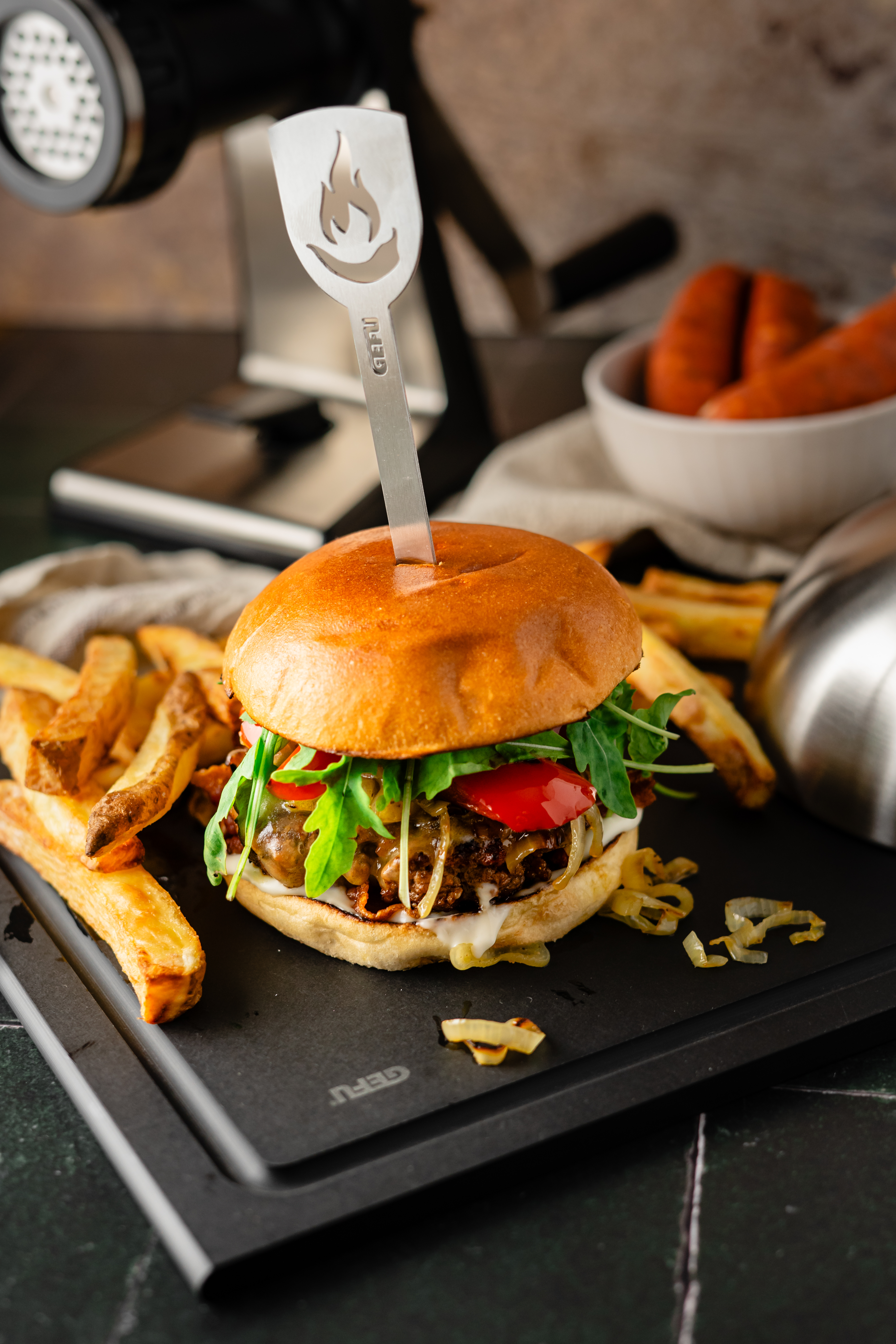 EM-Burger Spanischer Art Ein frisch zubereiteter Burger mit Salat und Paprika, serviert mit knusprigen Pommes auf einer schwarzen Servierplatte.