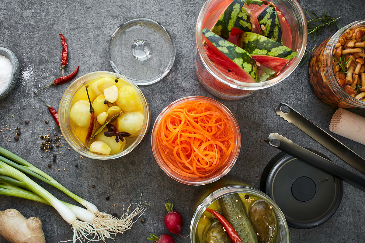 Verschiedene eingelegte Lebensmittel in Gläsern mit frischem Gemüse und Gewürzen auf einer Küchenarbeitsplatte Gläser mit eingelegten Zwiebeln, Karotten, Wassermelone, Gurken und Pilzen auf einer grauen Küchenplatte, umgeben von Frühlingszwiebeln, Chilischoten, Pfefferkörnern und Küchengeräten.
