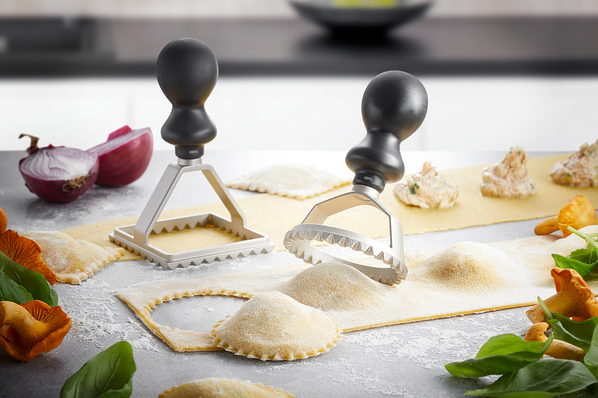 Zwei Metall-Ravioli-Ausstechformen auf einem Tisch mit Teig und frischen Zutaten wie Zwiebel, Pilzen, und Spinat