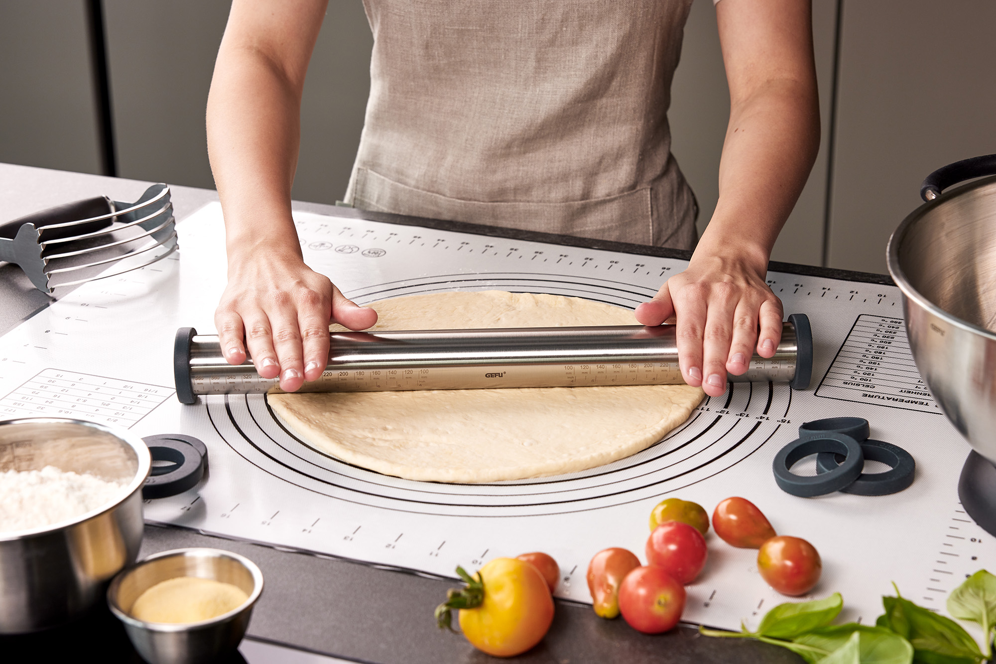 Teig wird mit einem Edelstahl-Teigroller ausgerollt Person rollt Teig mit Edelstahl-Teigroller auf einer Backmatte aus, umgeben von Mehl, Tomaten und Küchengeräten