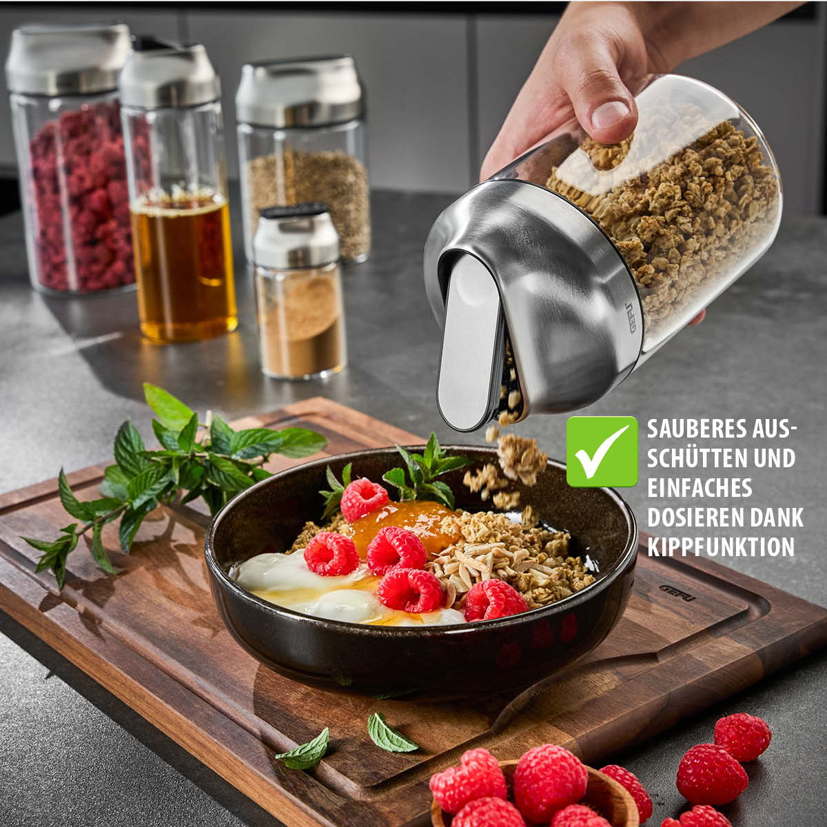 Vorratsbehälter beim Ausgießen von Müsli in eine Schüssel mit Joghurt und frischen Himbeeren, umgeben von weiteren Vorratsgläsern