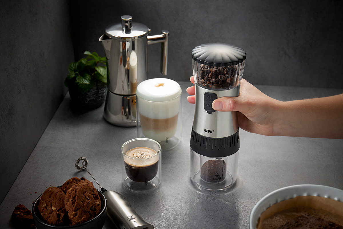 Elektrische Kaffeemühle im Einsatz neben Espressomaschine und Cappuccino Hand benutzt elektrische Kaffeemühle auf Küchentisch mit Espressomaschine, Cappuccino, Keksen und Pflanze