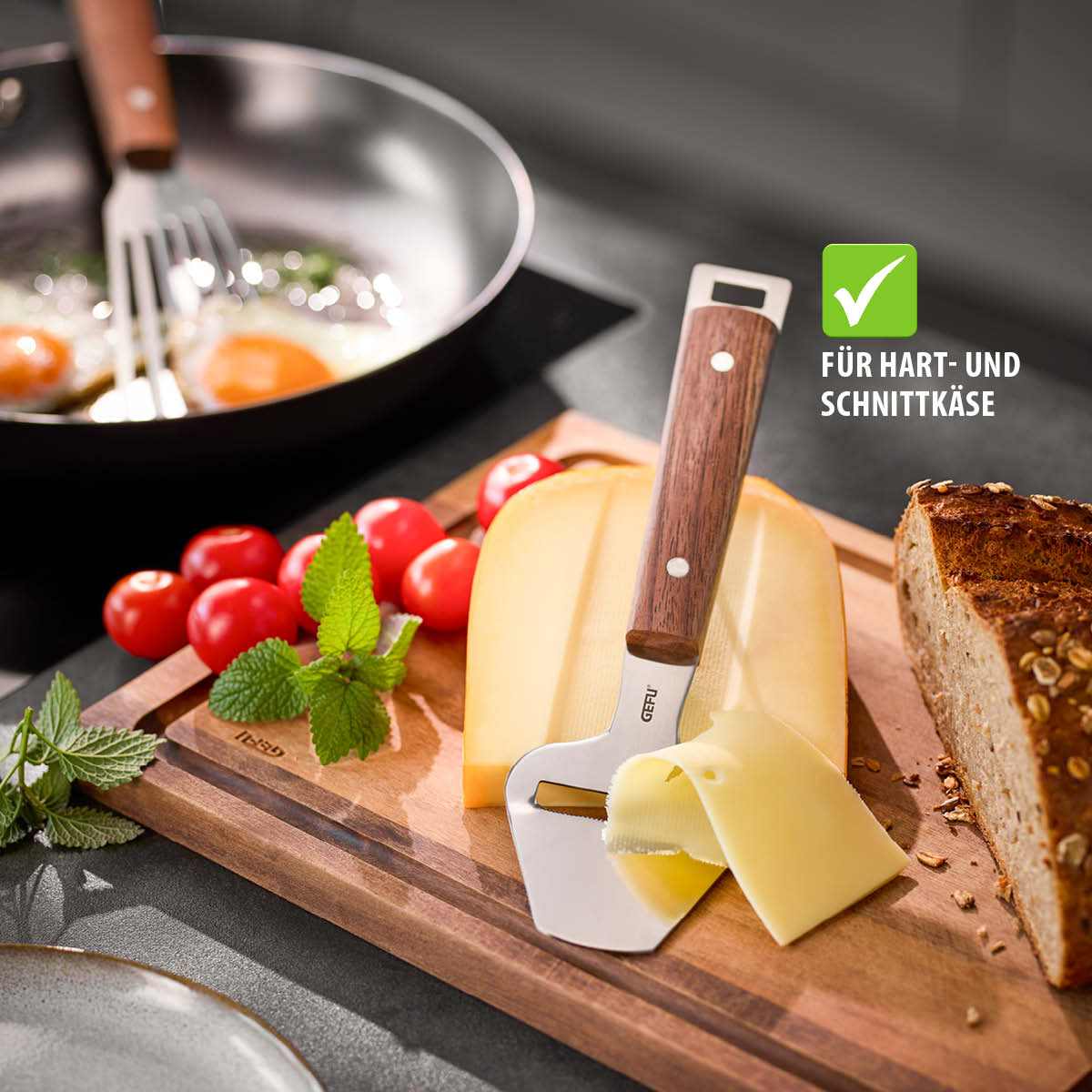 Ein Käsehobel mit Holzgriff liegt auf einem Schneidebrett neben einer Scheibe Käse, Tomaten und Brot