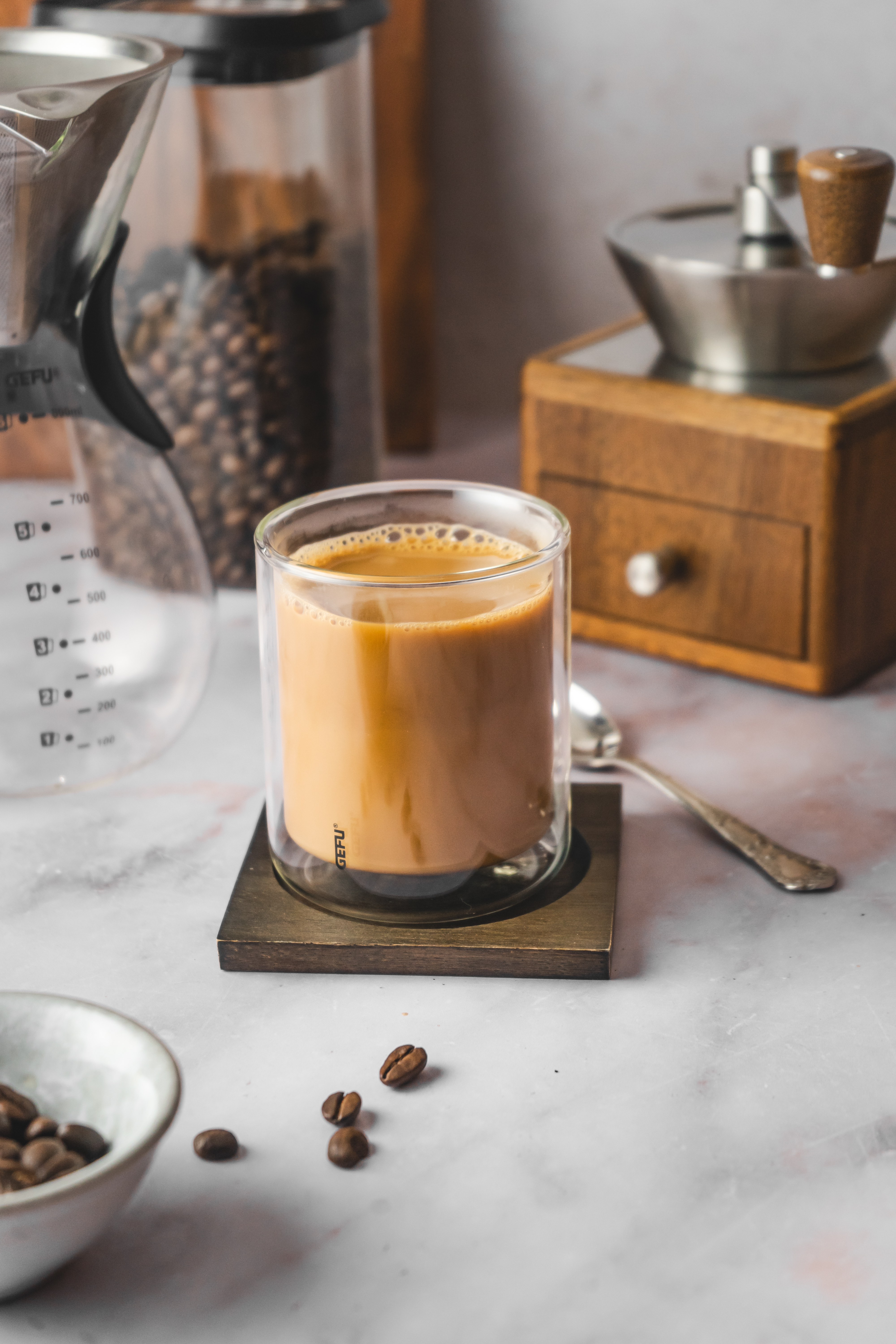 Filterkaffee Ein doppelwandiges Glas mit Kaffee, umgeben von einer Kaffeemühle, Kaffeebohnen und einem Kaffeefilter auf einem hellen Untergrund.