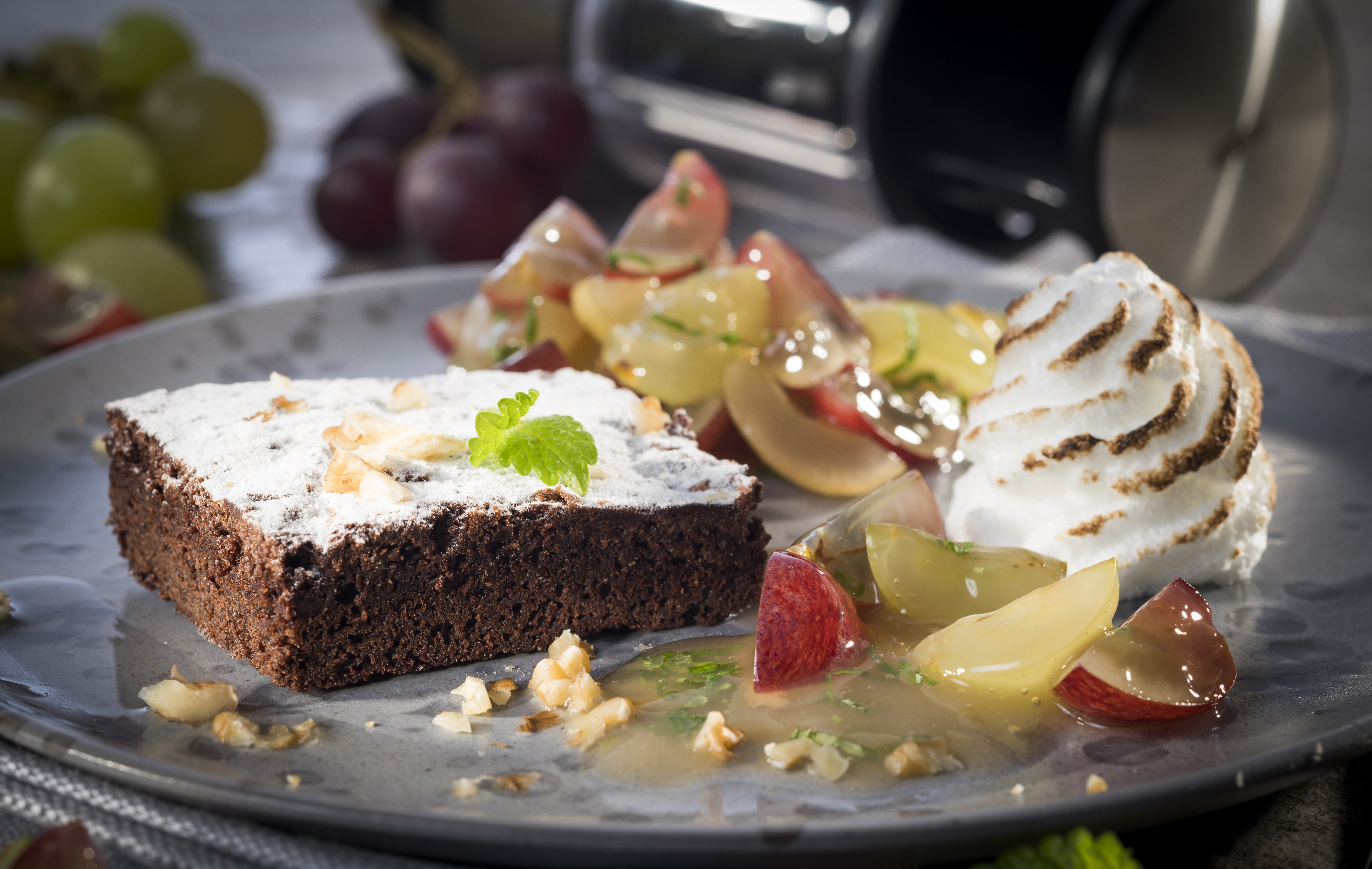 Schokoladen-Walnuss-Brownie mit Weintraubenragout und Baiser Ein Teller mit einem Stück Schokoladenkuchen, verziert mit Puderzucker und Nüssen, dazu frische Trauben und Sahne.