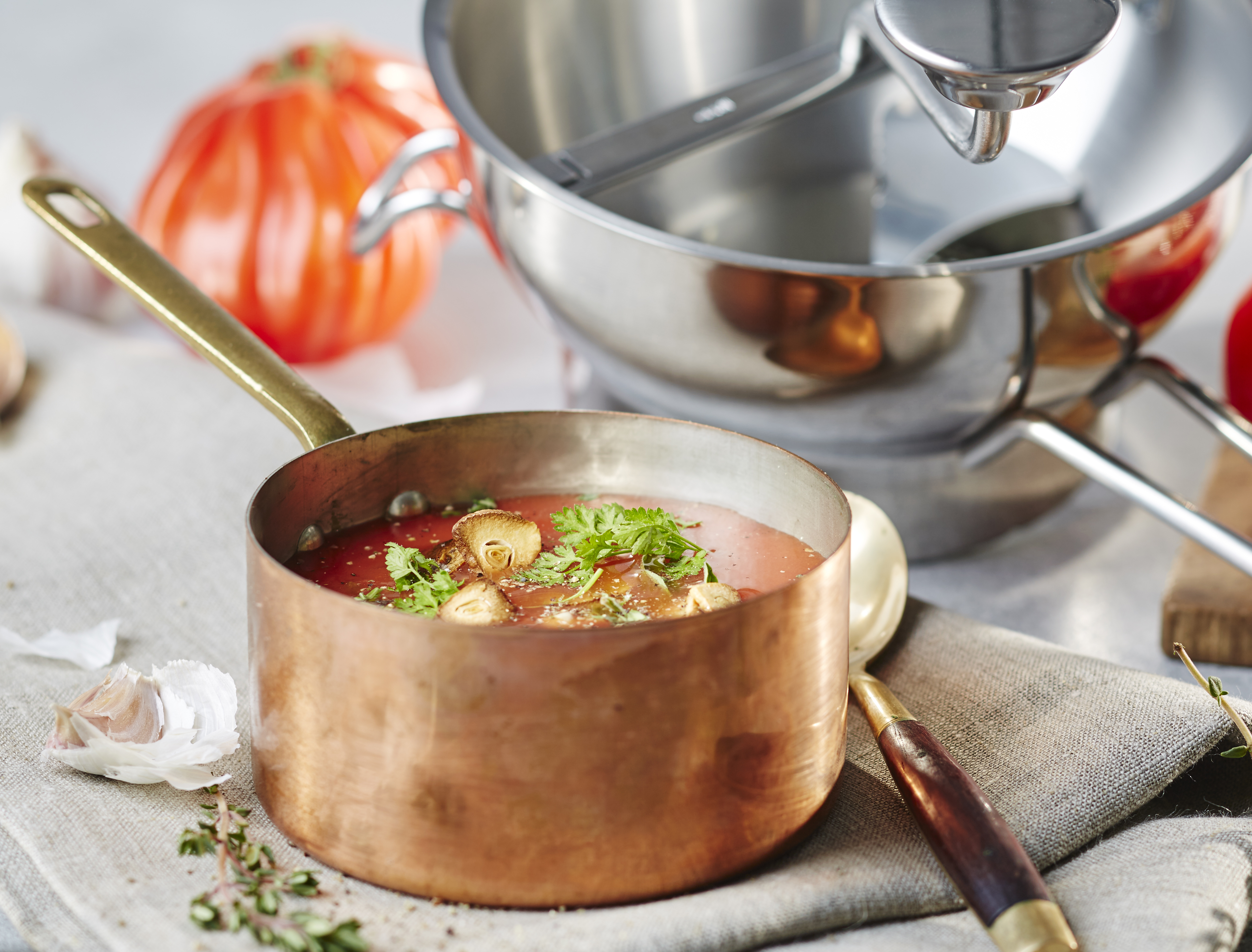Ofentomatensuppe mit Knoblauchchips Kupfertopf mit frisch zubereiteter Tomatensuppe, dekoriert mit Kräutern und Knoblauch, im Hintergrund ein Edelstahltopf und eine Tomate