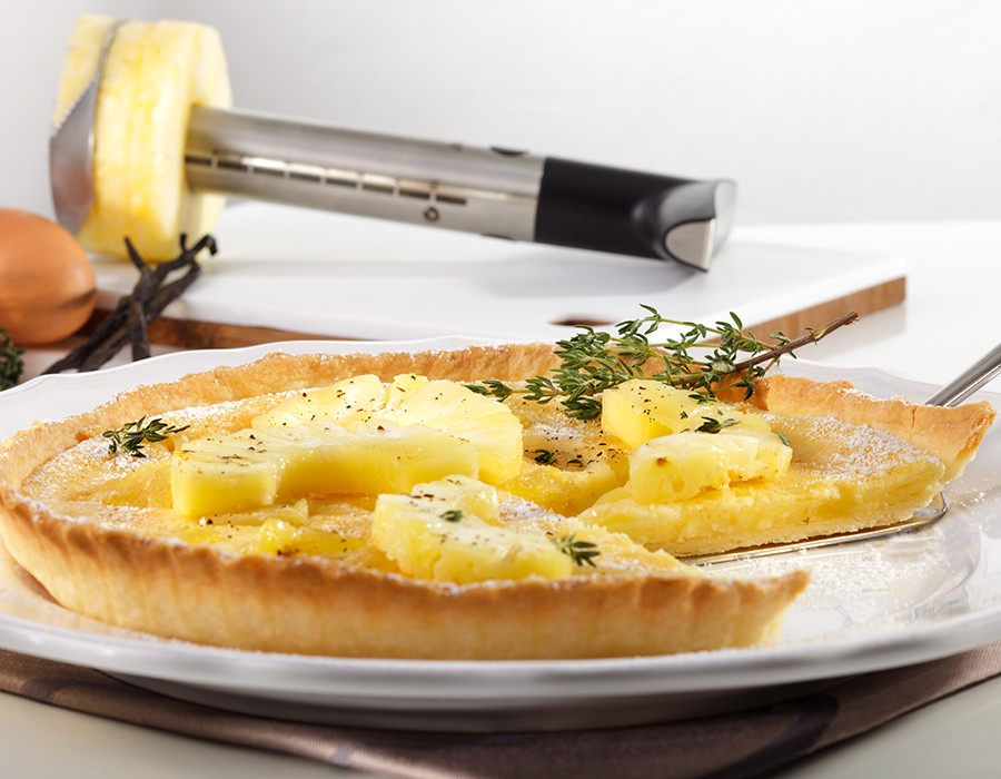Ananas-Tarte mit Mascarpone Eine frisch gebackene Tarte mit Ananas und Kräutern auf einem Teller, im Hintergrund ein Ananasschneider und Vanilleschoten.