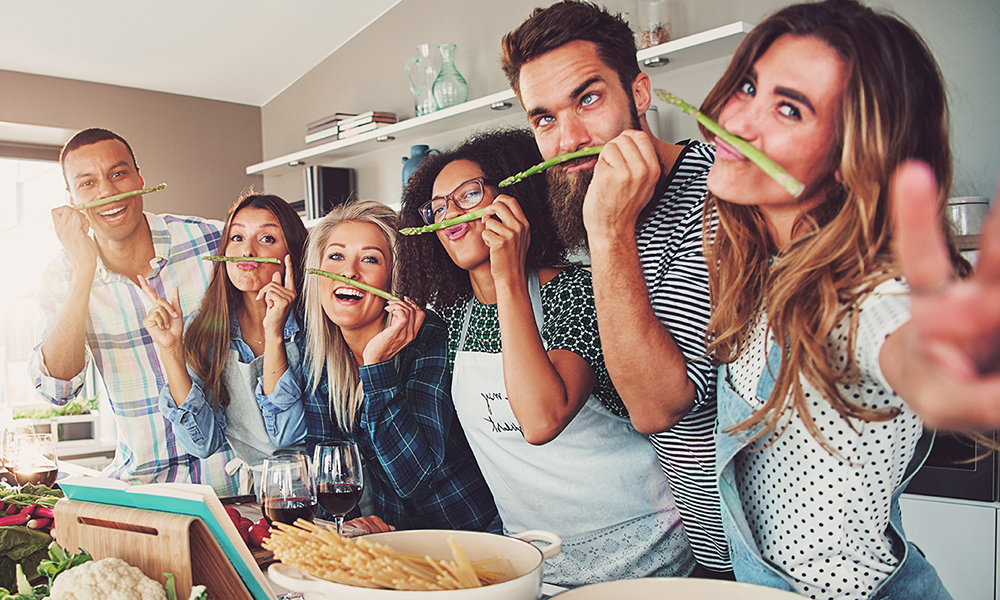 Freundesgruppe hat Spaß beim gemeinsamen Kochen in der Küche Sechs lachende Freunde in der Küche essen Spargel und genießen ein gemeinsames Kochabenteuer