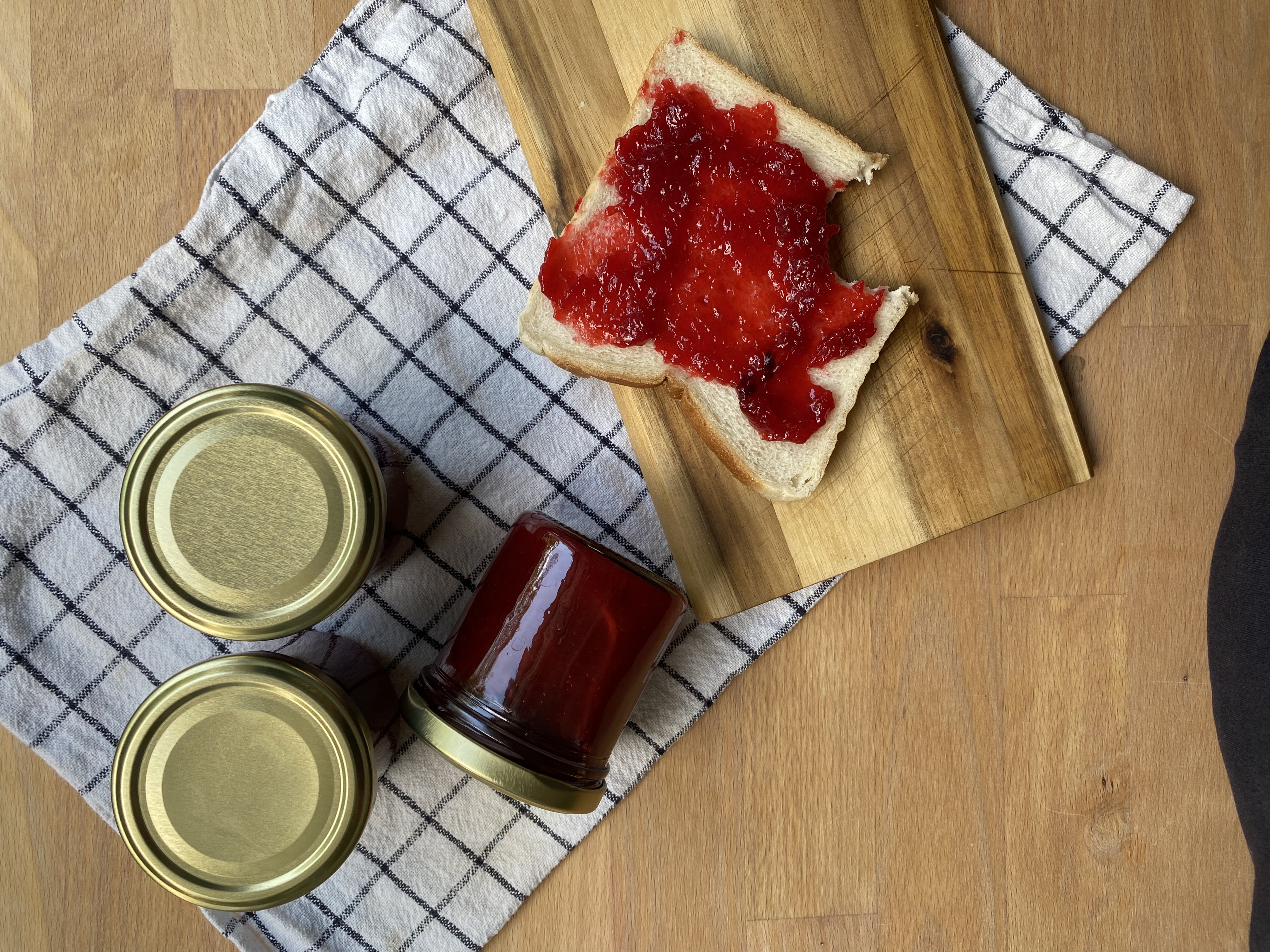 Himbeermarmelade Drei Gläser Erdbeermarmelade auf einem karierten Tuch und ein Toast mit Erdbeermarmelade auf einem Holzbrett