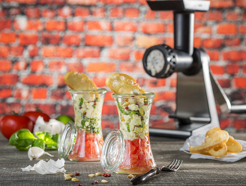 Seeteufeltatar auf Tomatenragout im Glas Zwei Gläser mit frischer Gemüsesalsa, garniert mit herzhaften Chips, im Vordergrund eines Zerkleinerers vor einer Ziegelwand.