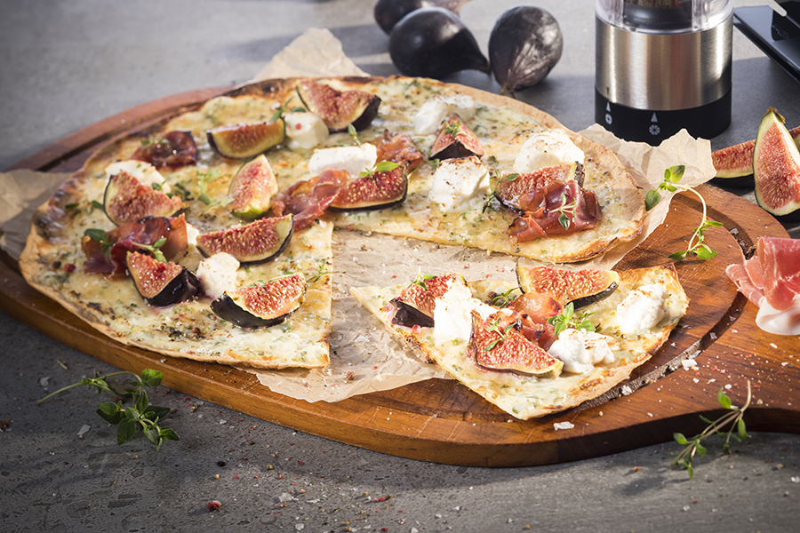Flammkuchen mit Feigen, Ziegenkäse, Thymianhonig und Parmaschinken Ein leckeres Feigen-Flammkuchen-Gericht auf einem Holzbrett serviert, garniert mit Schinken und Kräutern.