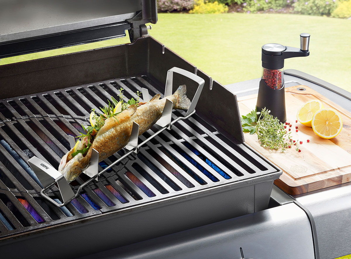 Fisch in Grillhalter auf einem Gasgrill, gewürzt mit Zitronenscheiben und Kräutern, daneben eine Pfeffermühle