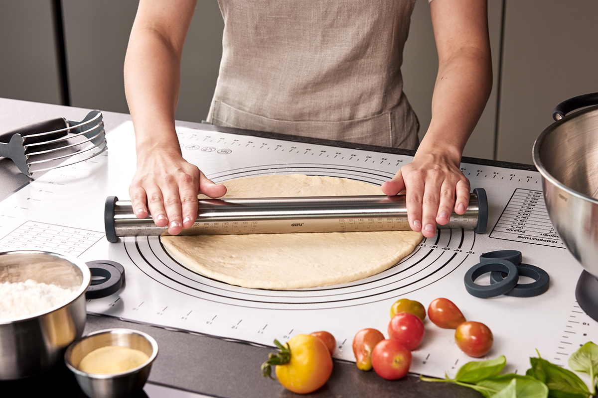 Teig ausrollen mit hochwertigem Edelstahl-Teigroller auf Antihaft-Backmatte Person rollt Teig mit einem Edelstahl-Teigroller auf einer Antihaft-Backmatte aus, umgeben von Schüsseln mit Mehl und Tomaten.