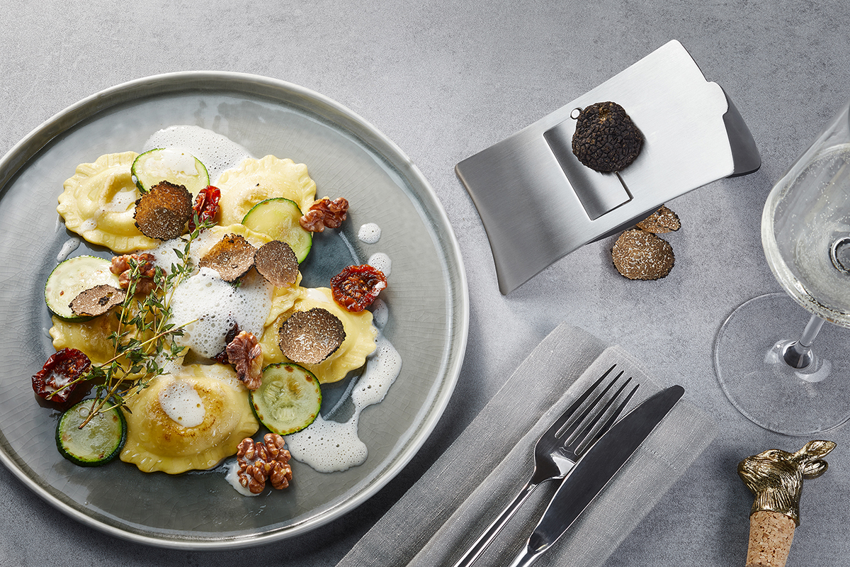 Ravioli mit Trüffelscheiben, Zucchini, getrockneten Tomaten und Walnüssen auf einem grauen Teller, daneben ein Trüffelschneider