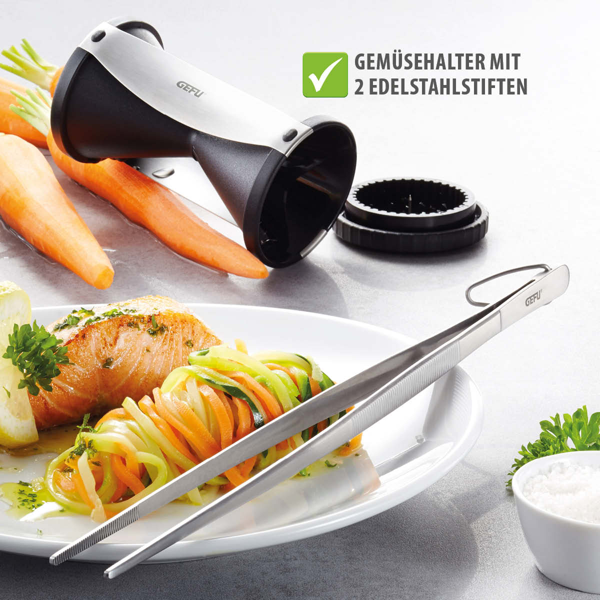 Spiralschneider für Gemüsenudeln mit Gemüsehalter und Edelstahlstiften Ein Spiralschneider neben einem Teller mit Gemüsespaghetti und Lachs. Enthält einen Gemüsehalter mit Edelstahlstiften.