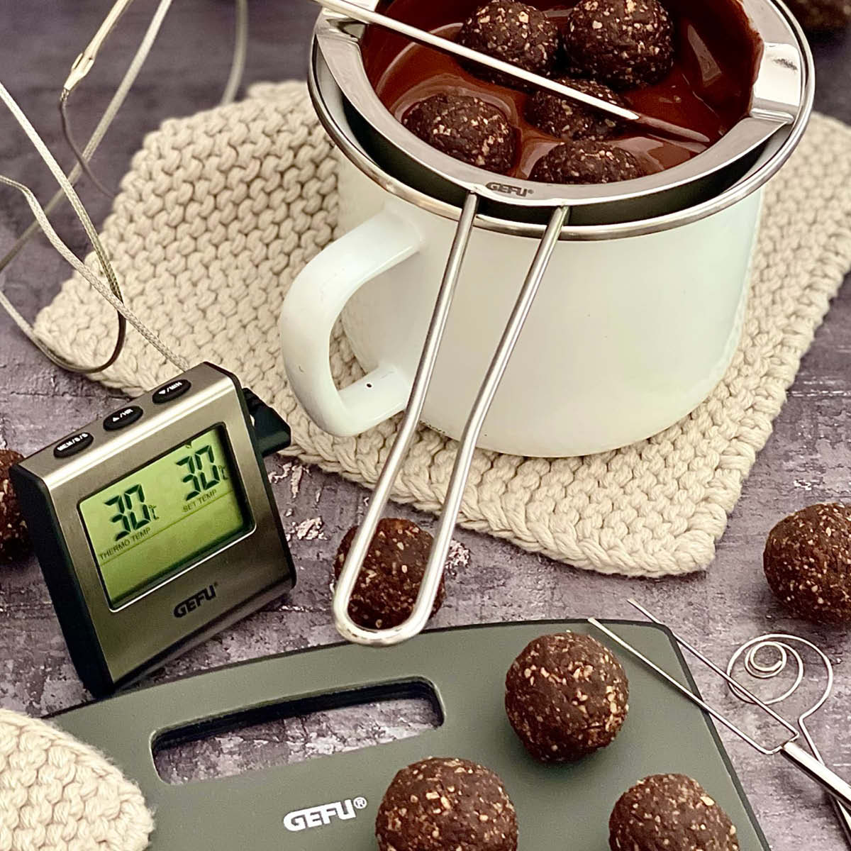 Schokoladenschmelzer mit Kugeln und digitalem Thermometer Ein weißer Becher mit Schokoladenschmelzer, umgeben von Schokoladenkugeln und einem digitalen Thermometer