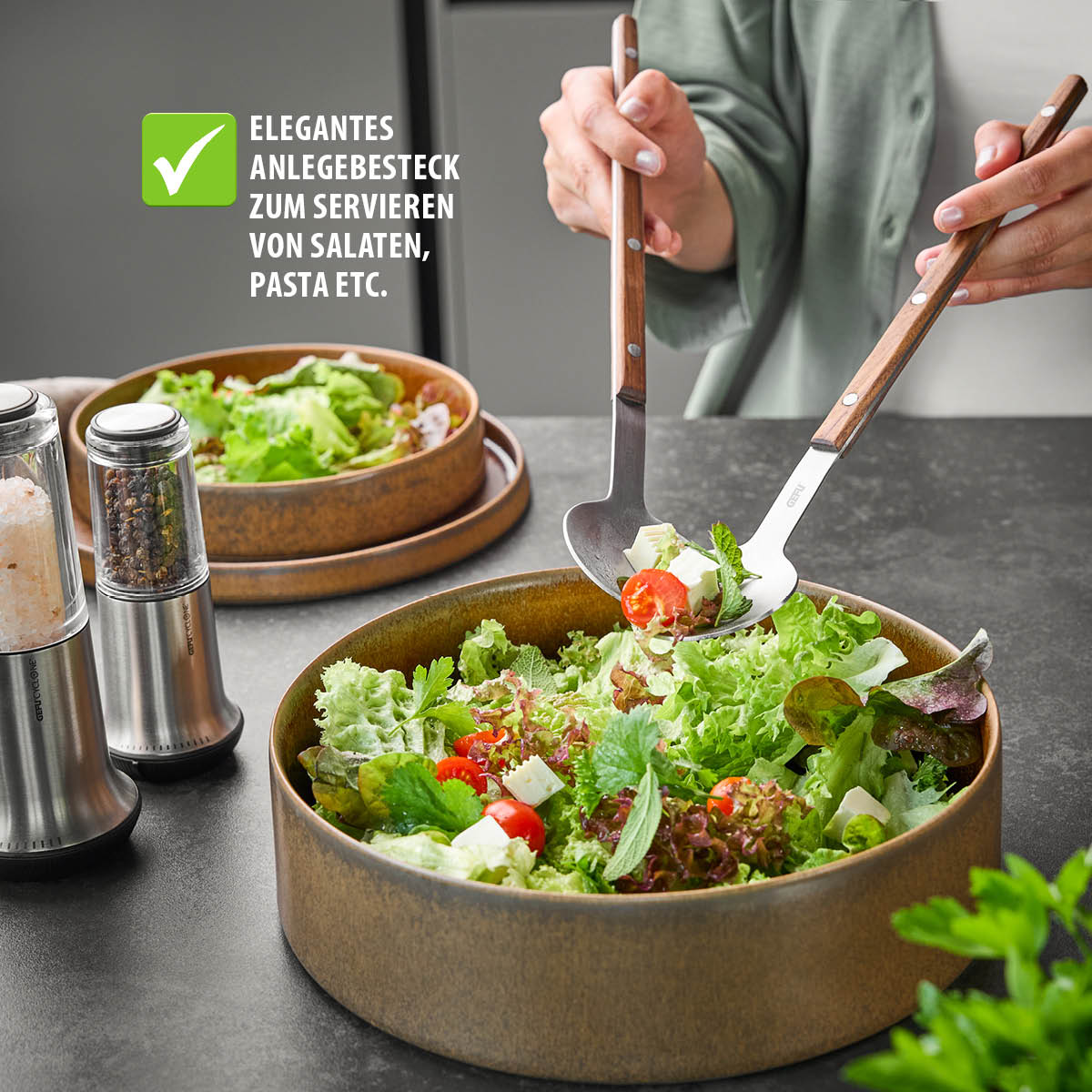 Nahaufnahme eines Salattellers mit Salatbesteck, das zum Servieren frischer, bunter Salate verwendet wird. Im Hintergrund ein Gefäß mit Gewürzmühlen.