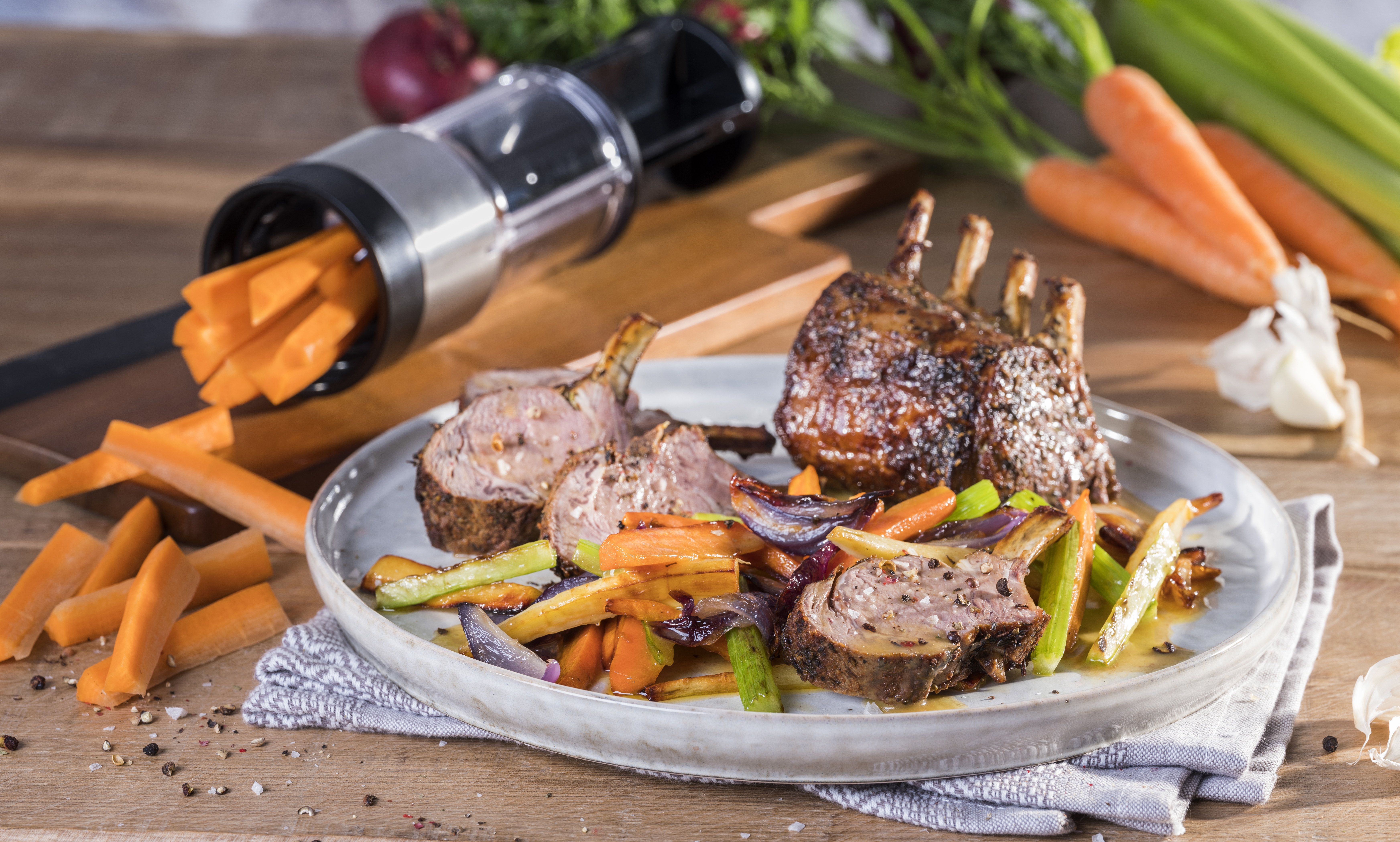 Kräuter-Lammkarree mit Marsala-Röstgemüse Ein Teller mit Lammkarree, gebratenem Gemüse und einem Gemüseschneider mit Karottenstiften im Hintergrund.