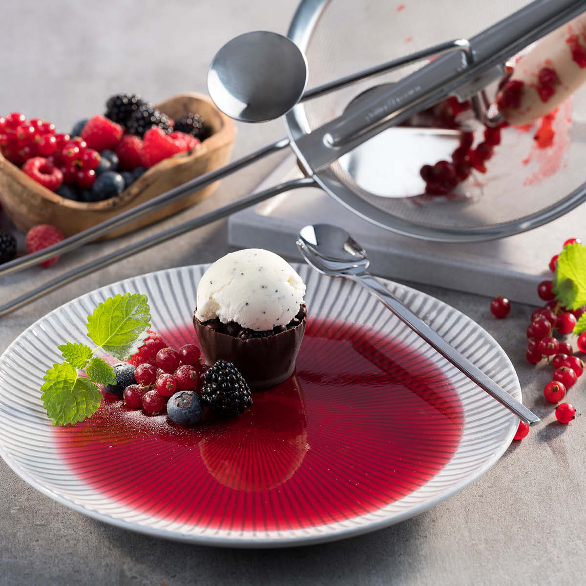 Ein Dessertteller mit Vanilleeis in einem Schokoladenbecher, umgeben von frischen Beeren und einer roten Beerensauce. Im Hintergrund liegt ein Löffel und weiteres Beerenobst in einer Holzschale.